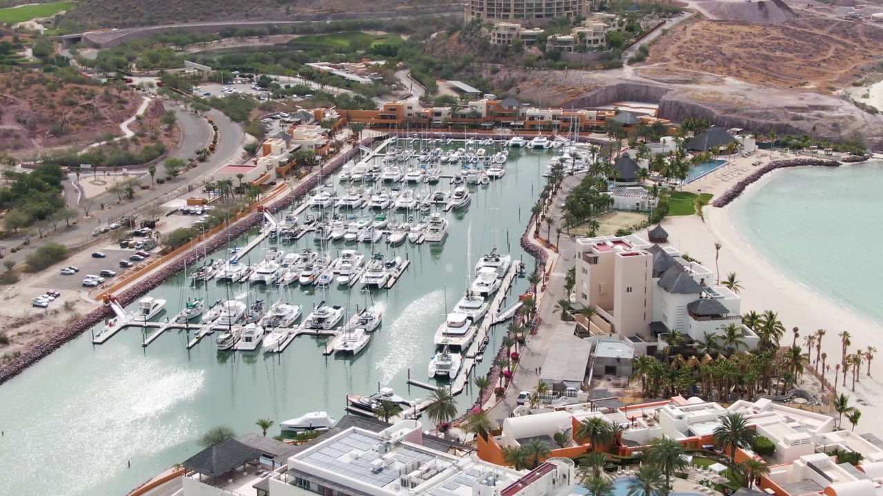 La Paz marina aerial with clustered boats and slips filling the center, a bright patch of rippled water in the lower left, a pale beach on the right, road traffic on the left, and no visible people.