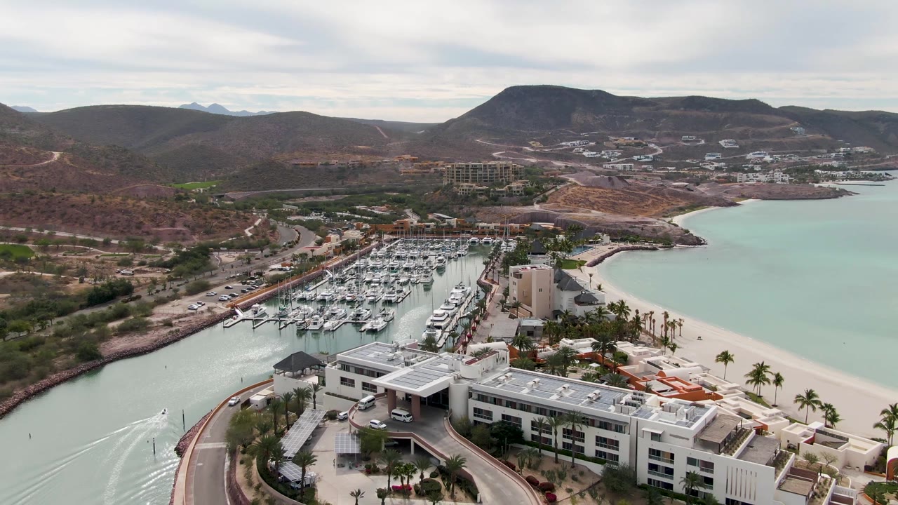 Aerial view in La Paz with the marina centered, pale blue water on the right, white waterfront buildings and palms below, dry hills beyond, and no visible people.