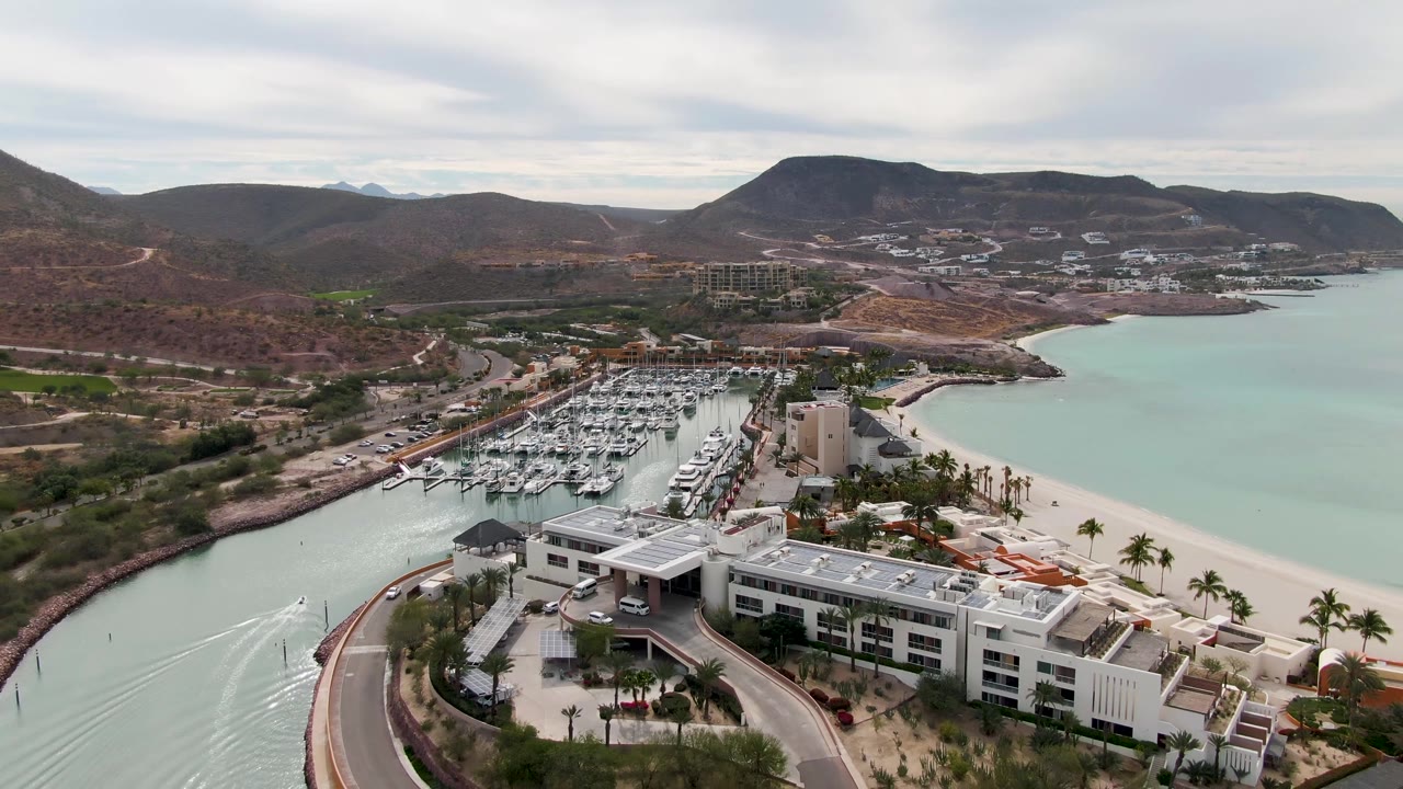 La Paz aerial frame with more of the curved road visible in the foreground, marina boats to the left, the sandy beach and calm water to the right, distant hills, and no visible people.
