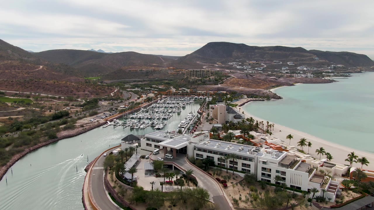 Aerial view with marina, curved road, beach, hills, and boat wake.