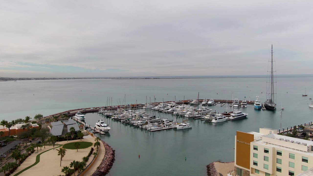 Aerial frame of a La Paz, Baja California Sur marina with docked boats, calm bay water, a pale waterfront building at lower right, palms along a path, gray sky, and no visible people.