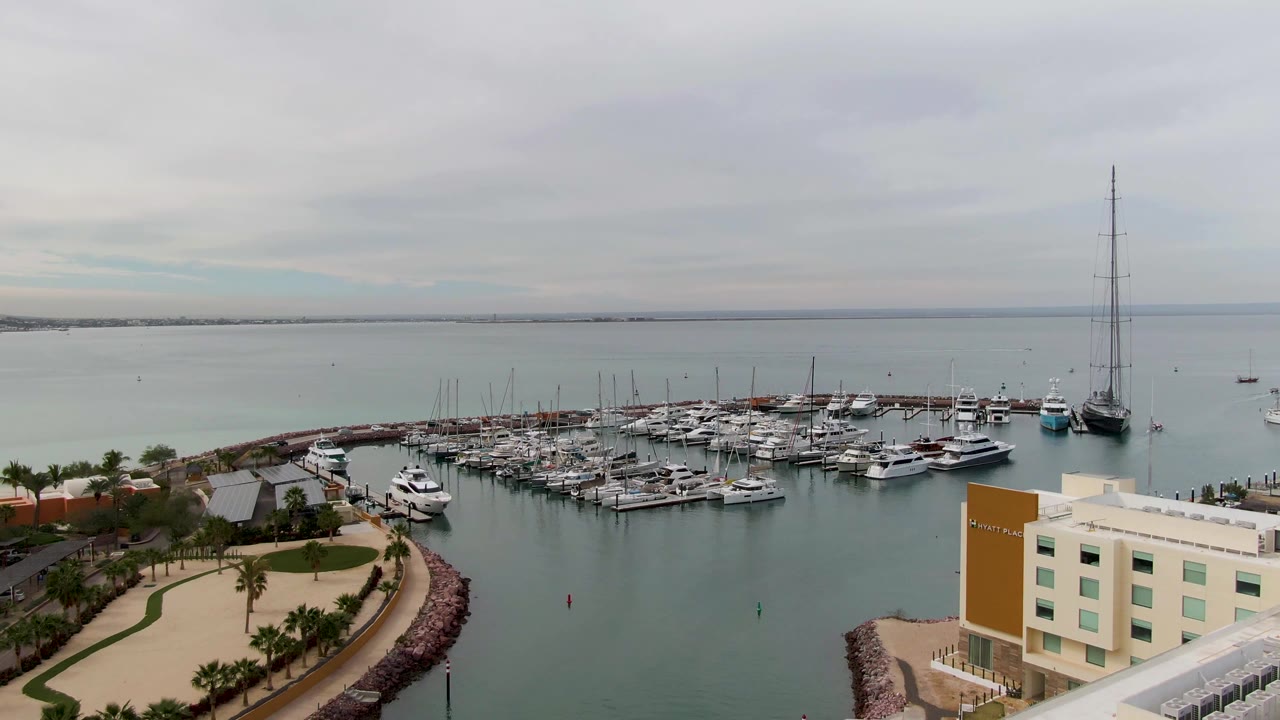 Aerial view in La Paz with the pale building more prominent in the lower right, curved palm-lined path below, marina docks across the middle, calm water beyond, and no visible people.