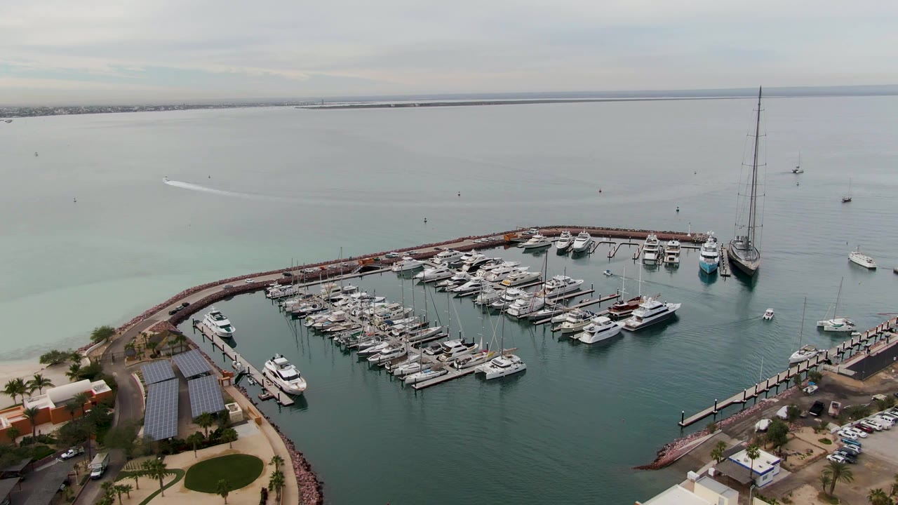 Opening aerial frame over a La Paz, Baja California Sur marina with clustered docked yachts, a curved breakwater, pale green coastal water, distant shoreline, and no visible people.