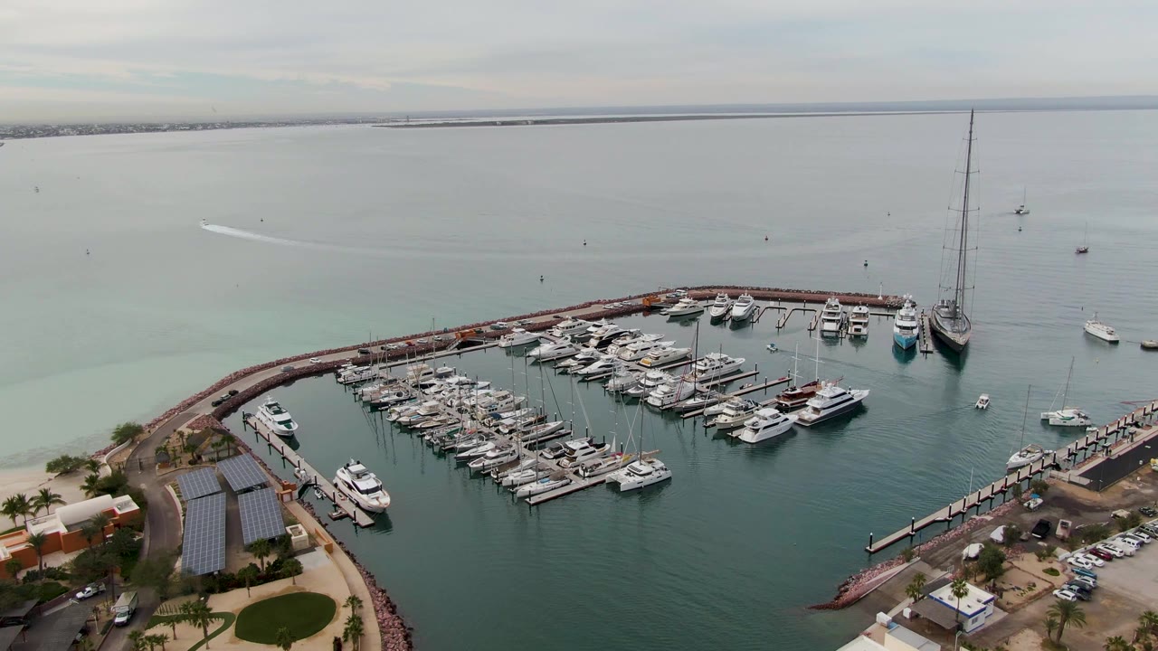 Aerial frame in La Paz, Baja California Sur showing the marina from slightly farther along the harbor, with dock rows, a long pier at right, offshore boats, muted clouds, and no visible people.