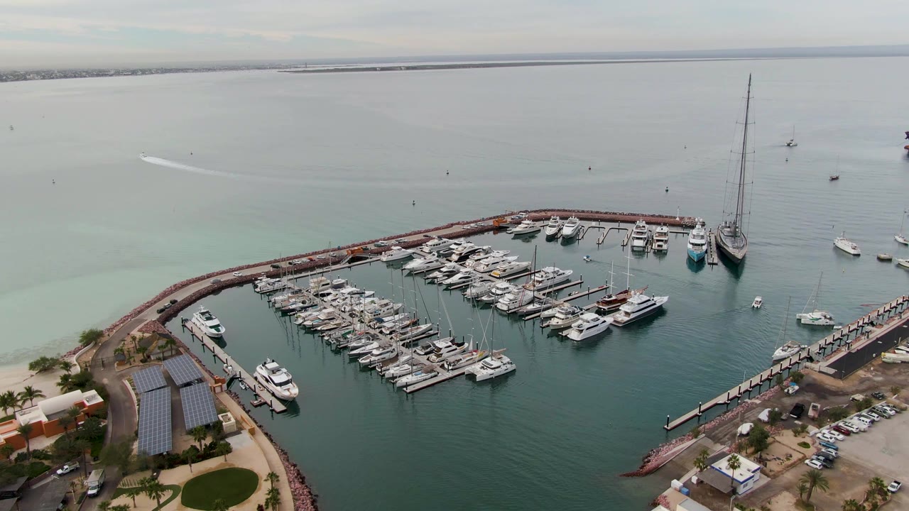 Aerial view across a marina, shoreline road, parked cars, and calm water.
