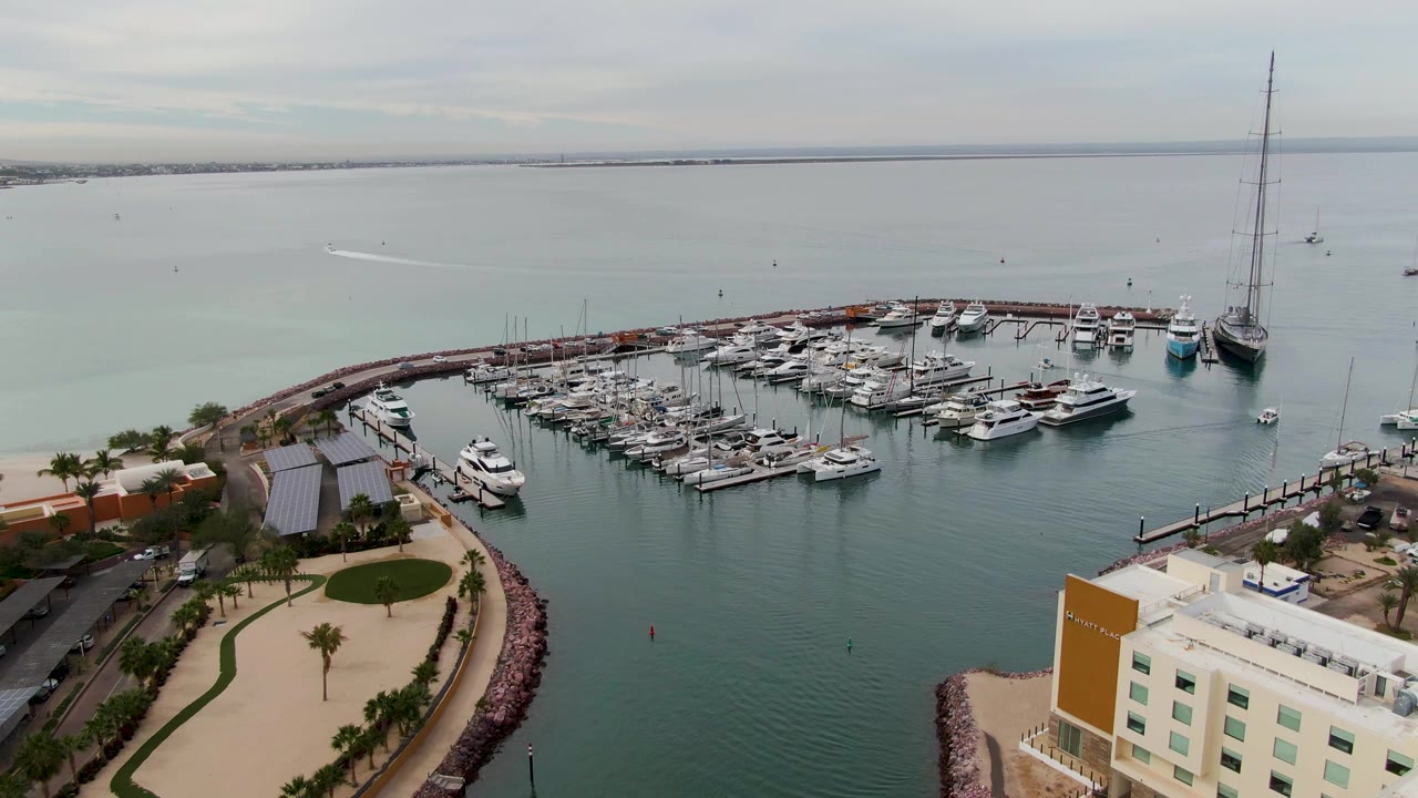 Aerial frame over a La Paz marina with rows of docked boats, a curved breakwater, pale green water, waterfront buildings, and no visible people.