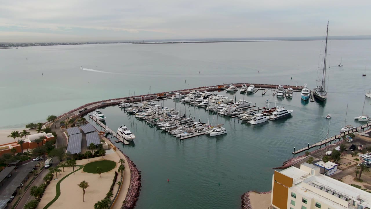 Aerial frame in La Paz showing docked boats inside the marina, open bay water beyond the breakwater, a small wake line in the distance, and no visible people.