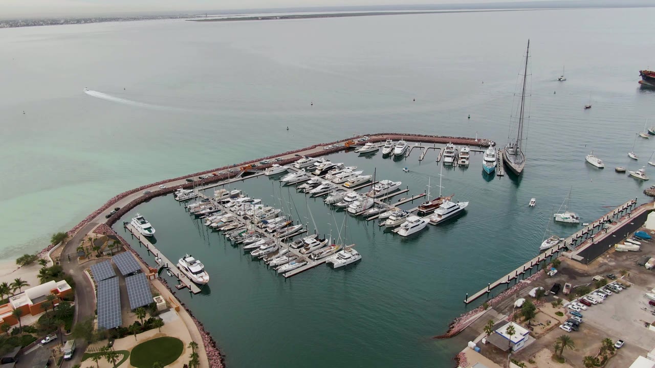 Aerial frame over a La Paz, Baja California Sur marina showing rows of docked boats inside a curved breakwater, shoreline buildings at the lower edge, calm water beyond, and no visible people.