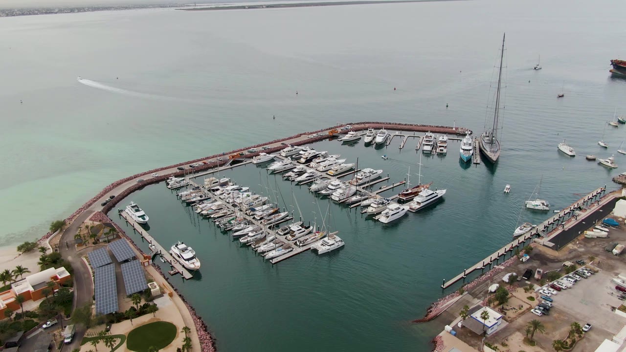 Aerial frame over La Paz showing the marina basin filled with white boats, pale turquoise shallows to the left, deeper water inside the harbor, shoreline structures, parked cars, and no visible people.