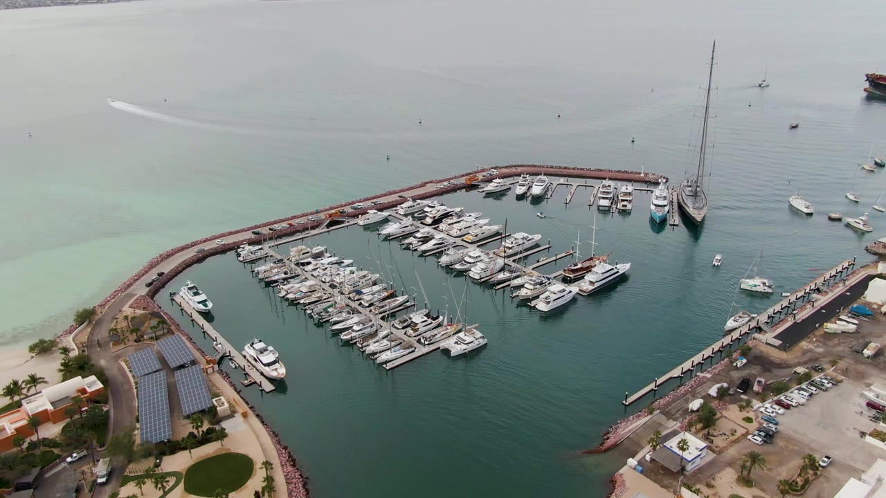La Paz aerial frame with docked boats grouped inside the breakwater, a tall sailboat mast near the right side, small boats on open water beyond the harbor, shoreline buildings, and no visible people.