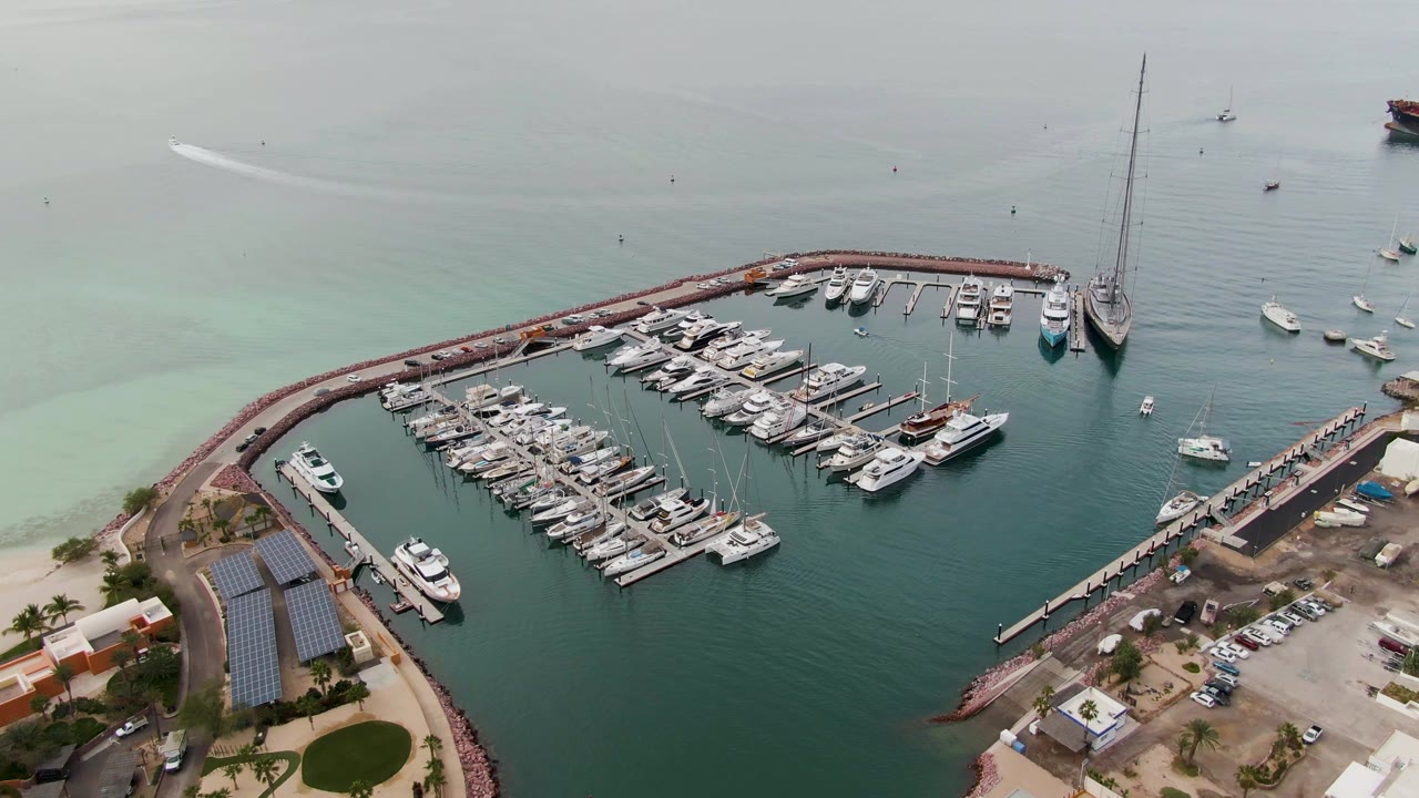 Aerial view of the marina, docked boats, breakwater, shoreline buildings, and calm coastal water.