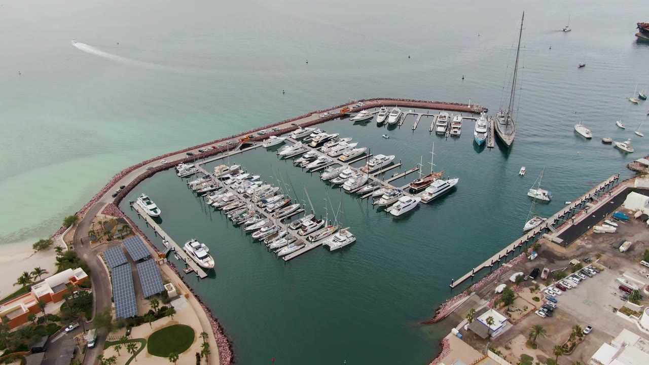 Aerial view over a La Paz, Baja California Sur marina with rows of white boats inside a curved breakwater, pale turquoise water outside, shoreline buildings below, and no visible people.