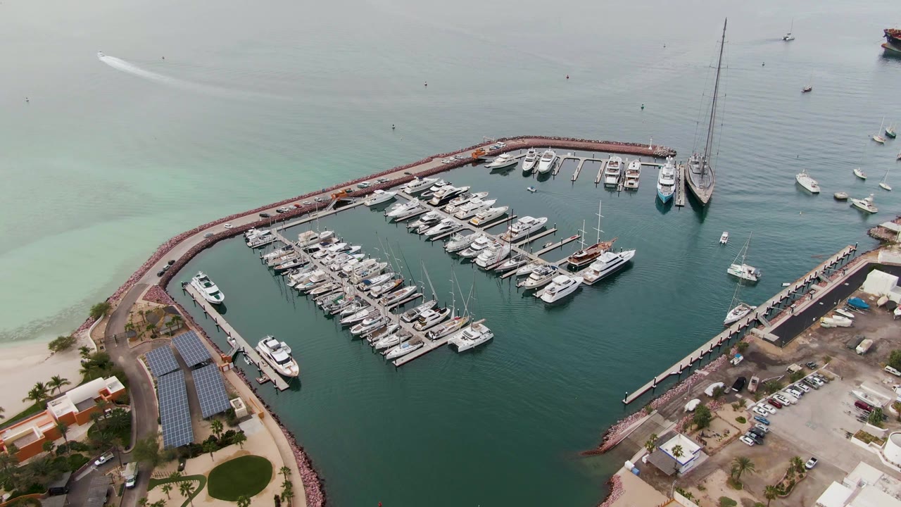 High aerial frame in La Paz, Baja California Sur showing docked yachts clustered inside the marina, a long pier to the right, scattered boats offshore, and no visible people.