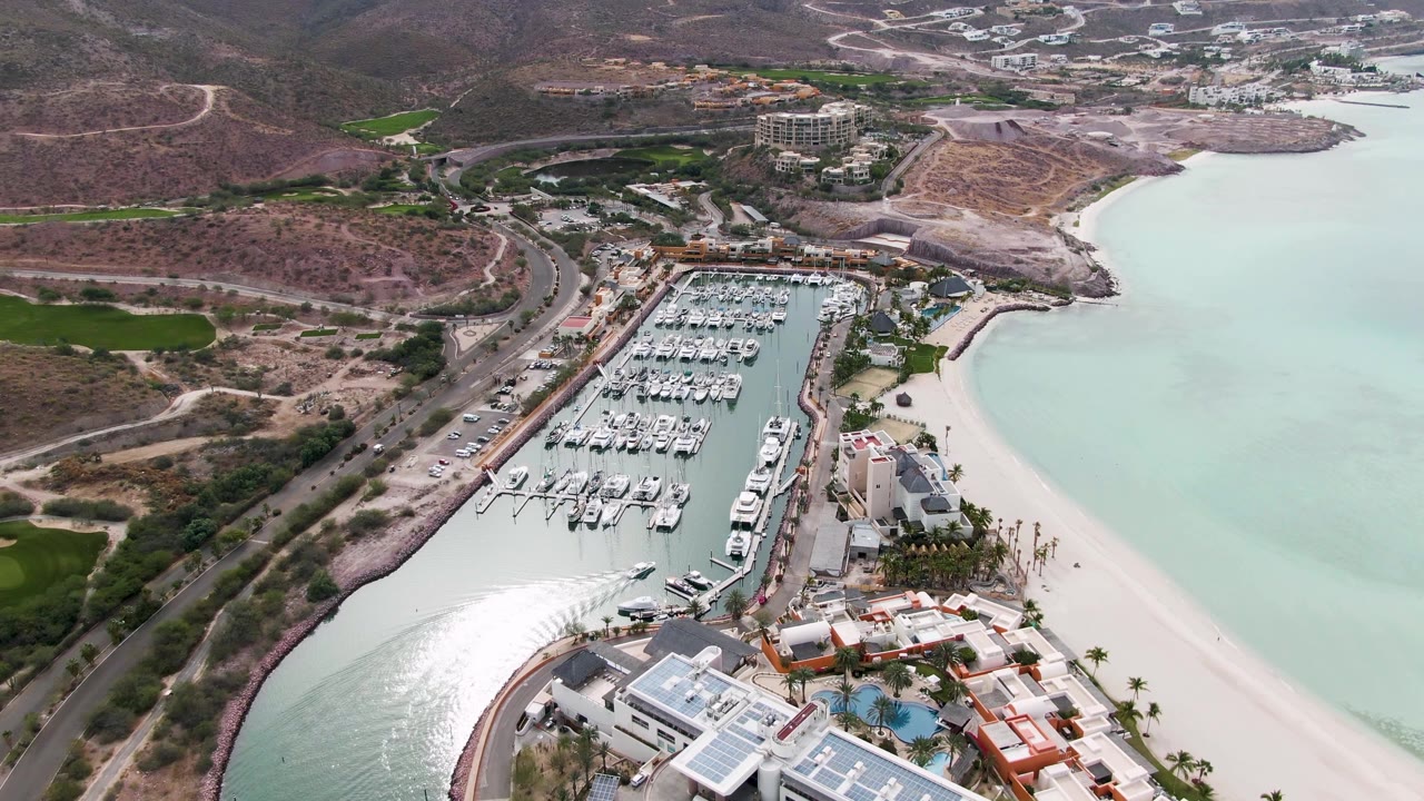 At two seconds, the La Paz marina remains in a wide aerial view with boat rows, pale sand, light green-blue water, hillside roads, buildings, and no visible people.