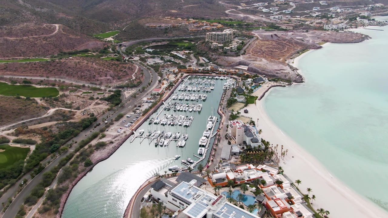 Aerial view of a coastal marina with boats, beach, water reflections, roads, and hills.