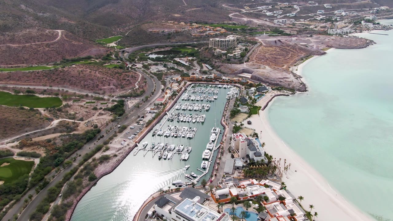 One second into the La Paz aerial, the marina remains centered with white boats in slips, a bright beach at right, a curved road at left, and no visible people.