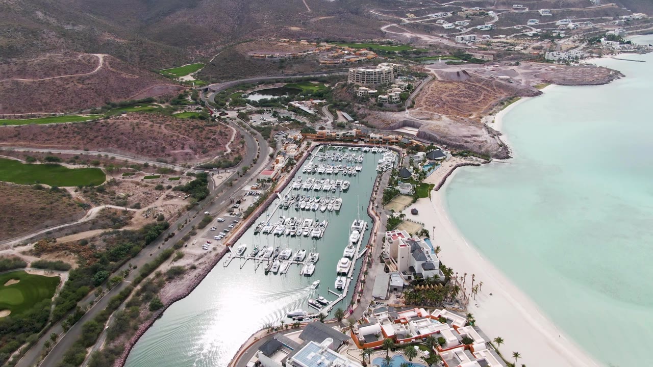 Aerial marina scene with bright water glare, boats, road, beach, buildings, and hills.
