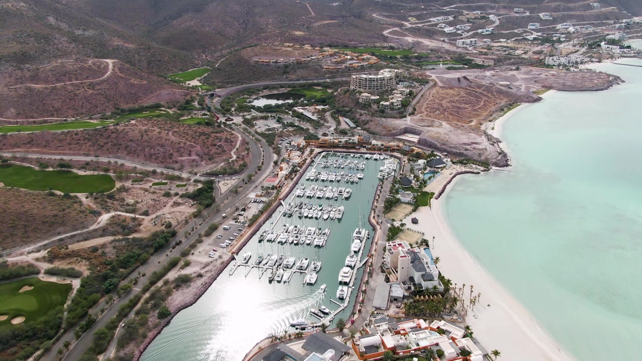 Opening aerial frame over La Paz, Baja California Sur, showing docked boats in a marina, pale beach water, a coastal road, green areas, dry hills, and no visible people.