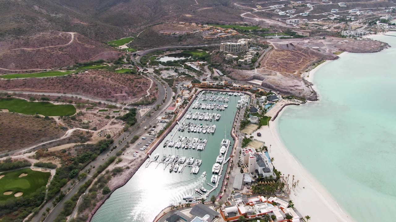 At 0:02, the aerial view keeps the La Paz marina centered, with green course-like areas on the left, dry hills behind, pale ocean water on the right, and no visible people.