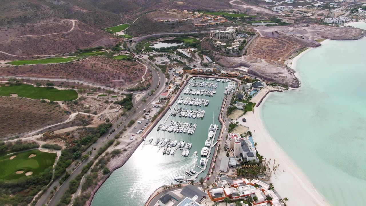 Aerial frame over La Paz, Baja California Sur showing a boat-filled marina, coastal road, pale beach, dry hills, green golf-course patches, and no visible people.