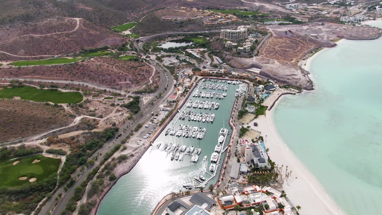 Second aerial frame over La Paz shows the marina basin and rows of docked boats with bright reflection on the lower harbor water and no visible people.