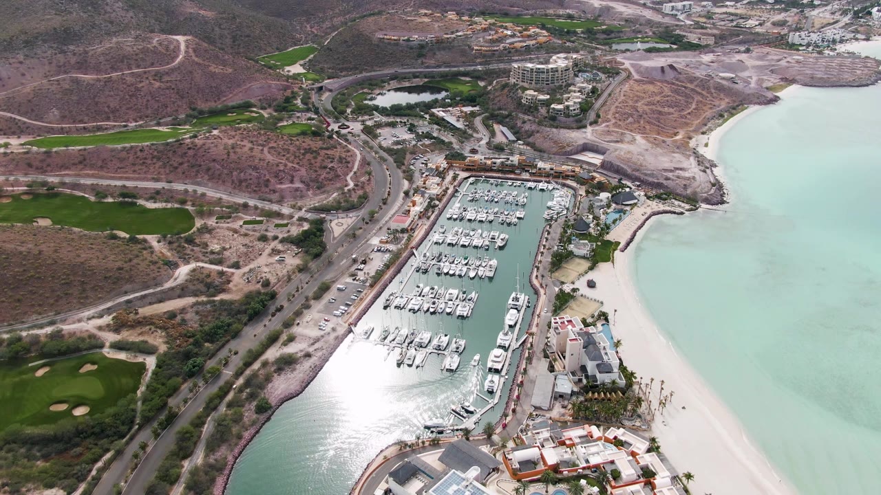 Aerial frame in La Paz with the marina centered, pale shoreline to the right, road and dry hillside to the left, green fairways in the background, and no visible people.