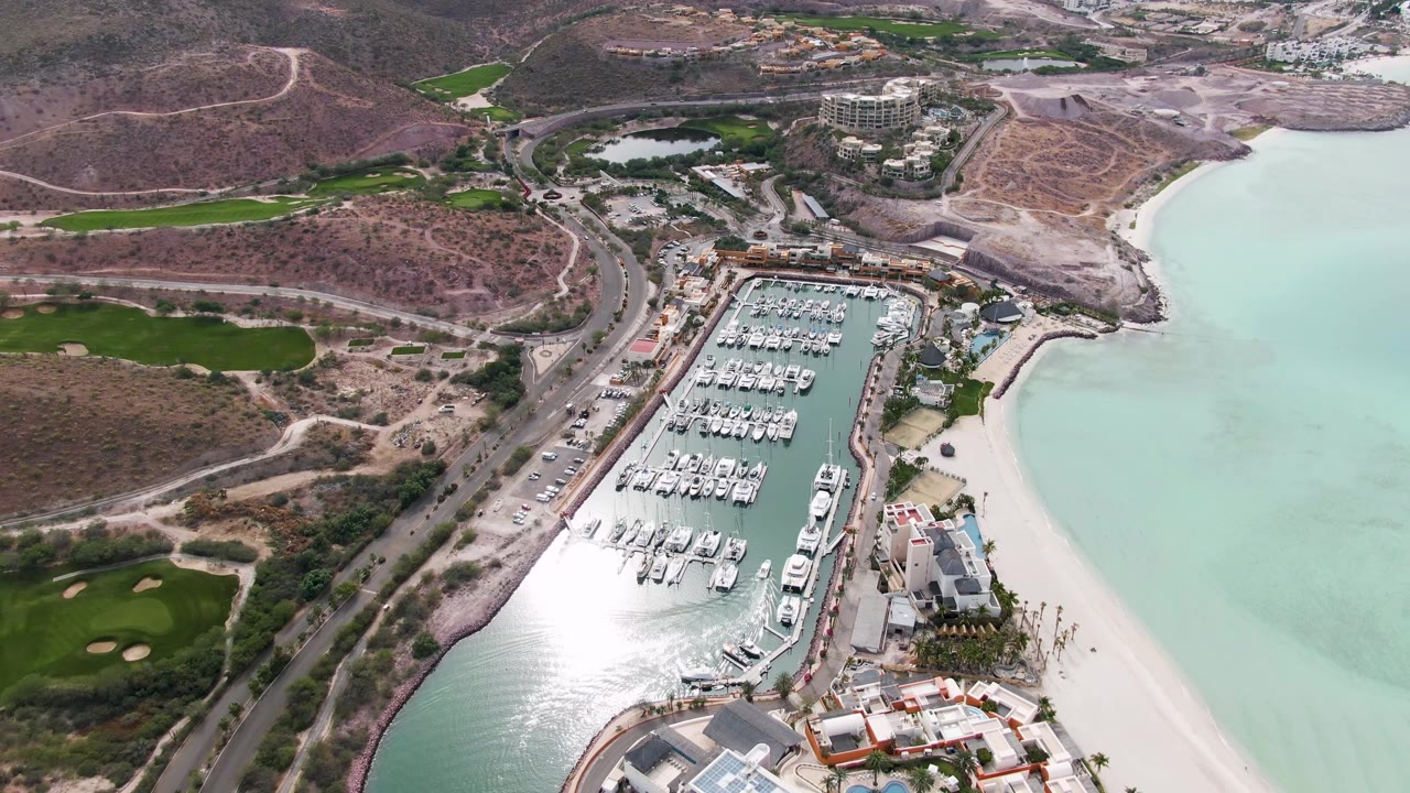 Aerial marina scene with boats, road traffic, beach, water reflection, and surrounding hills.