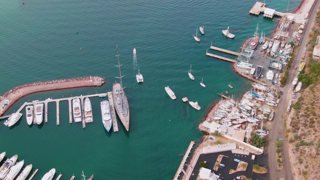 Opening aerial frame over a La Paz, Baja California Sur marina, showing docked white yachts, sailboats, a curved rock breakwater, boatyard edges, turquoise water, and no visible people.