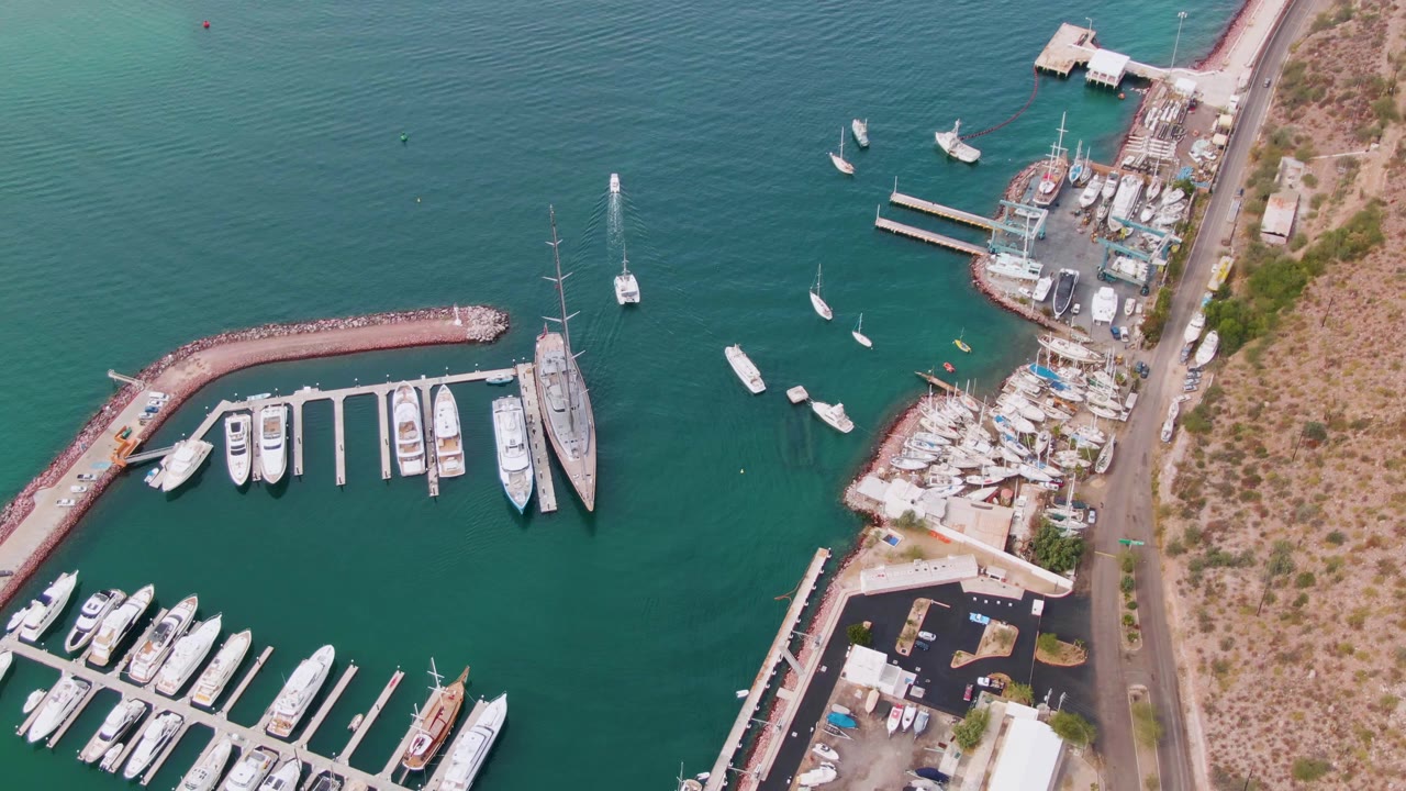High aerial frame over a La Paz, Baja California Sur marina with turquoise water, docked boats, a curved breakwater, shoreline road, dry land, and no visible people.