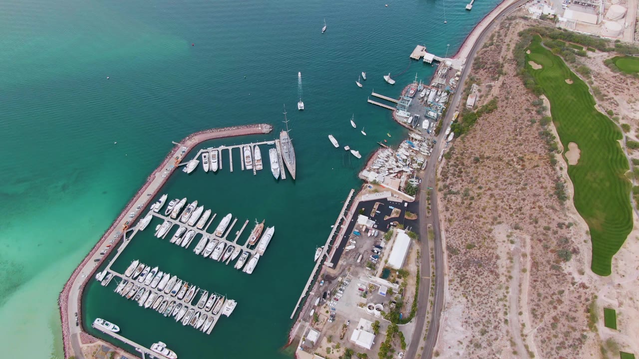 Opening overhead view of a La Paz, Baja California Sur marina with docked boats, a curved breakwater, turquoise shallows, a shoreline road, dry land, green grounds, and no visible people.