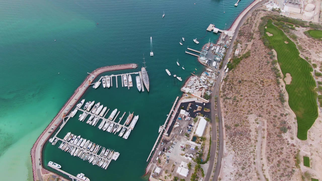One second into the clip, the La Paz marina remains centered with rows of white boats, small vessels in open water, dry shoreline lots, a road, green grounds, and no visible people.