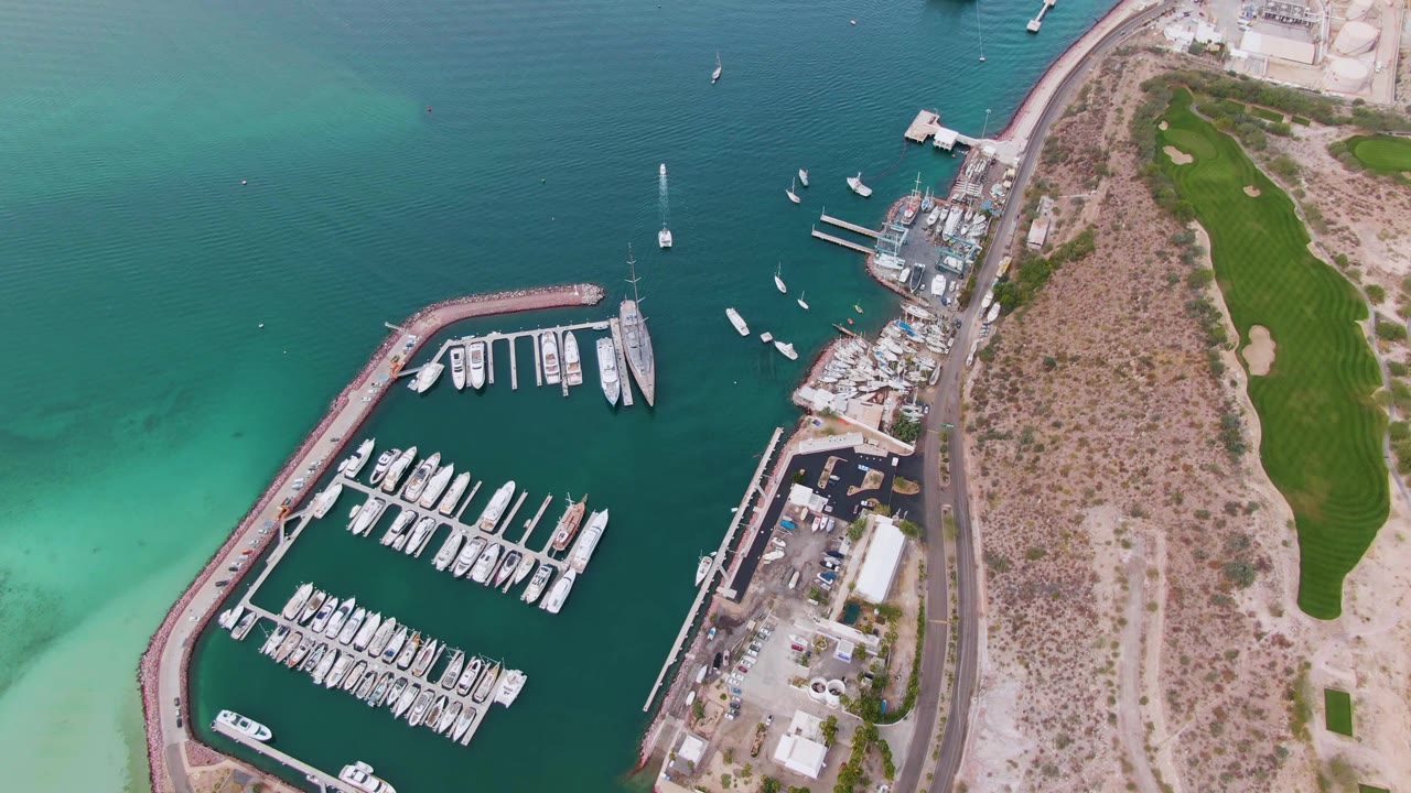 At 0:02, the aerial frame shows the La Paz marina basin and curved seawall, with turquoise shallows to the left, darker open water above, shoreline road and dry scrubland to the right, and no visible people.
