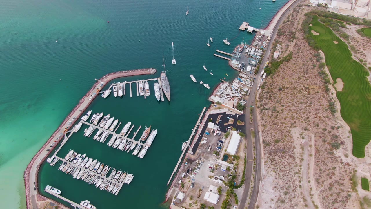 Aerial view of a marina, docked boats, turquoise water, road, and green land.