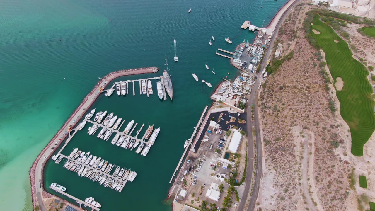 Drone-style aerial frame in La Paz showing docked yachts and slips, a small white vessel near the marina entrance, coastal road infrastructure, dry shoreline terrain, green field edges, and no visible people.