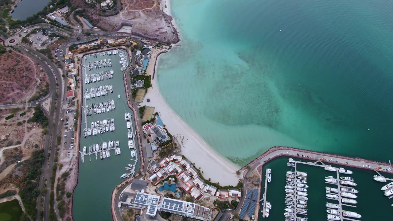 Overhead view of a La Paz marina with docked boats, a pale beach, curved breakwater, turquoise shallows, darker open water, and no visible people at the start of the clip.