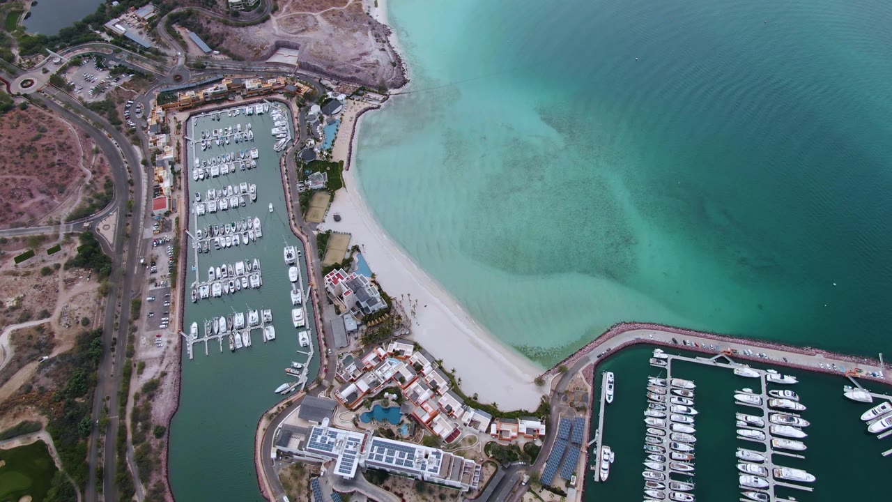 Aerial frame over La Paz showing the boat slips, curved shoreline, pale sand, and a soft ripple pattern spreading across the turquoise water, with no visible people.