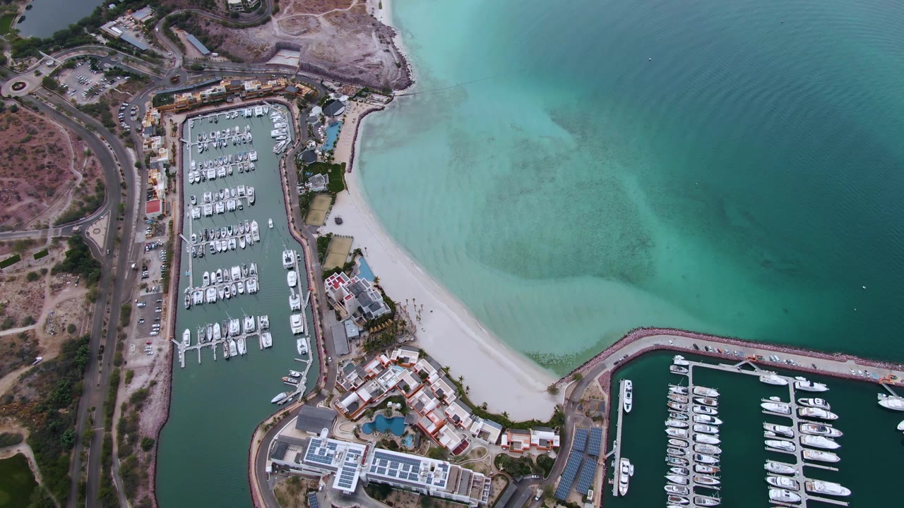 Opening aerial frame over La Paz, Baja California Sur showing docked boats in a marina, a pale sandy beach, shoreline buildings, a left-side road, and turquoise water with no visible people.