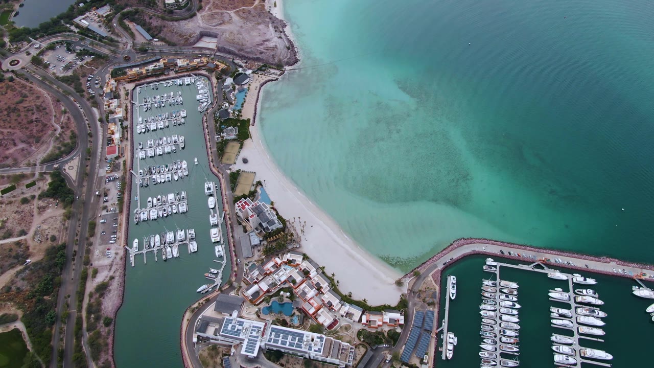 Second aerial frame in La Paz, Baja California Sur with the marina basin left of center, a curved sand strip below it, darker open water at right, and no visible people.