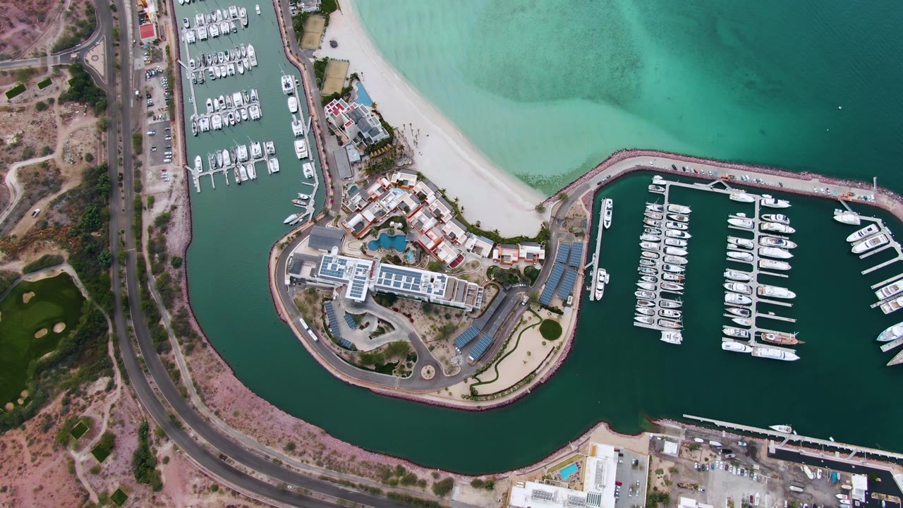 Overhead view of a La Paz, Baja California Sur marina with docked boats, a curved breakwater, a sandy beach, a coastal road at left, turquoise shallows at top, and no visible people.