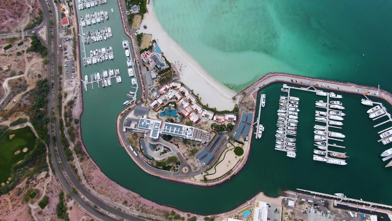 Drone view of marina docks, boats, curved breakwater, beach, road, and turquoise bay water.