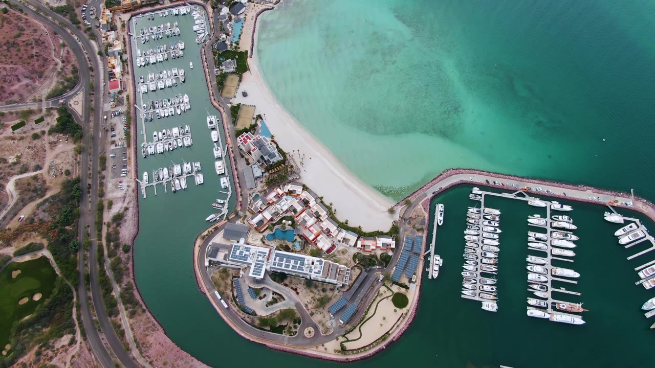 Opening overhead view of a La Paz, Baja California Sur marina with docked boats, a sandy beach, roads, green-blue water, and no visible people.