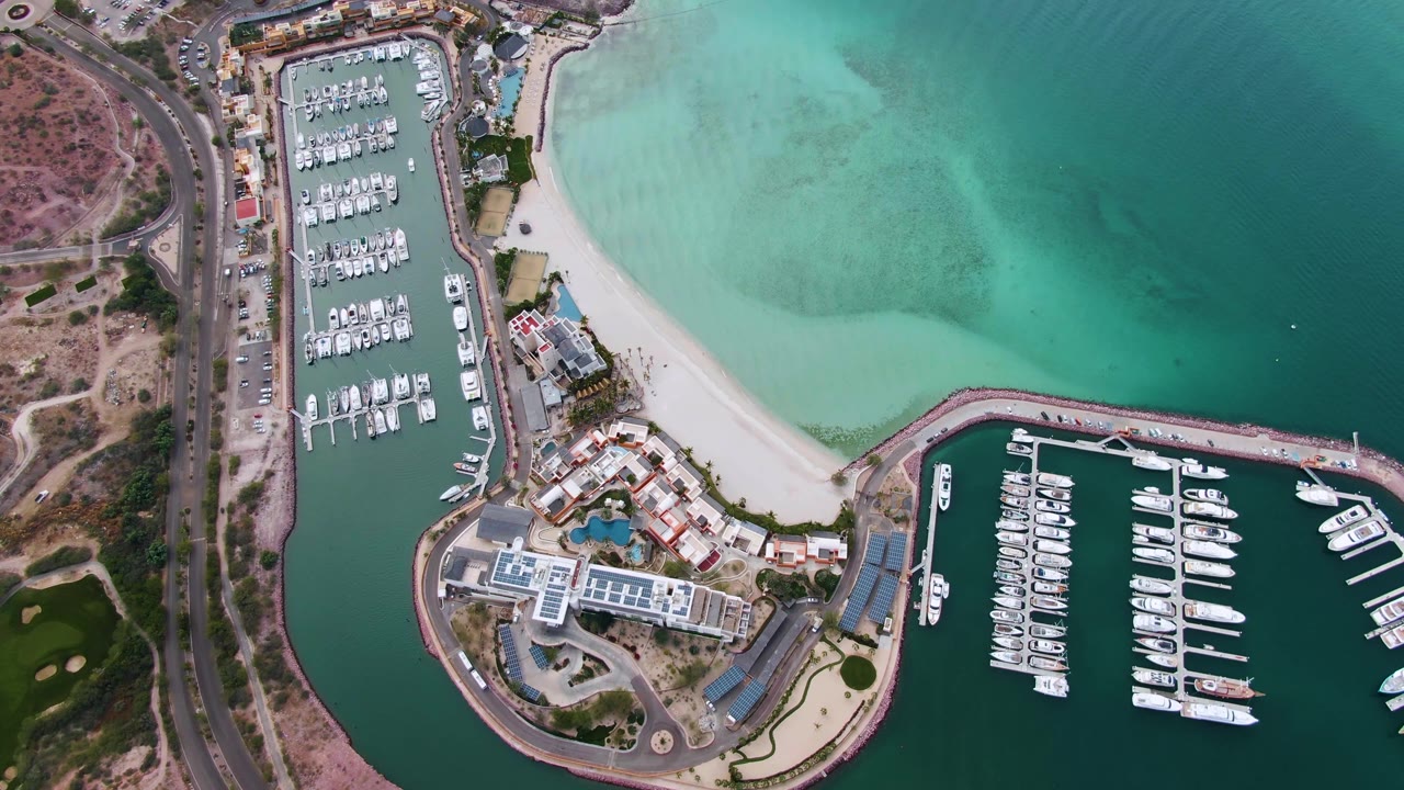 High aerial frame over La Paz showing the left marina packed with boats, the curved sandy beach, the lower harbor basin, roadside vehicles, and no visible people.