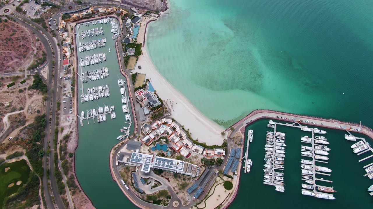 Overhead La Paz marina frame with boat rows in both basins, a pale sand shoreline, bright turquoise shallows, roadside traffic areas, and no visible people.