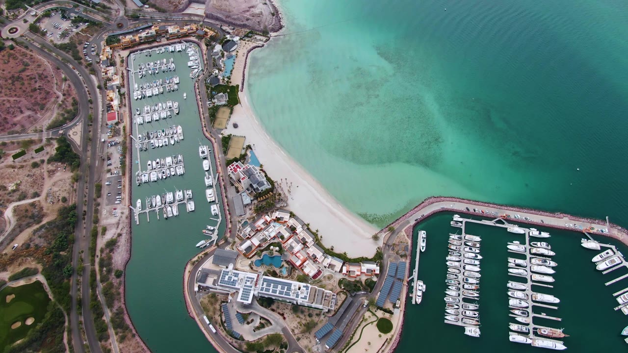 Mid-clip overhead view in La Paz with the road and dry terrain along the left edge, marina docks below, beach through the center, turquoise sea above, and no visible people.