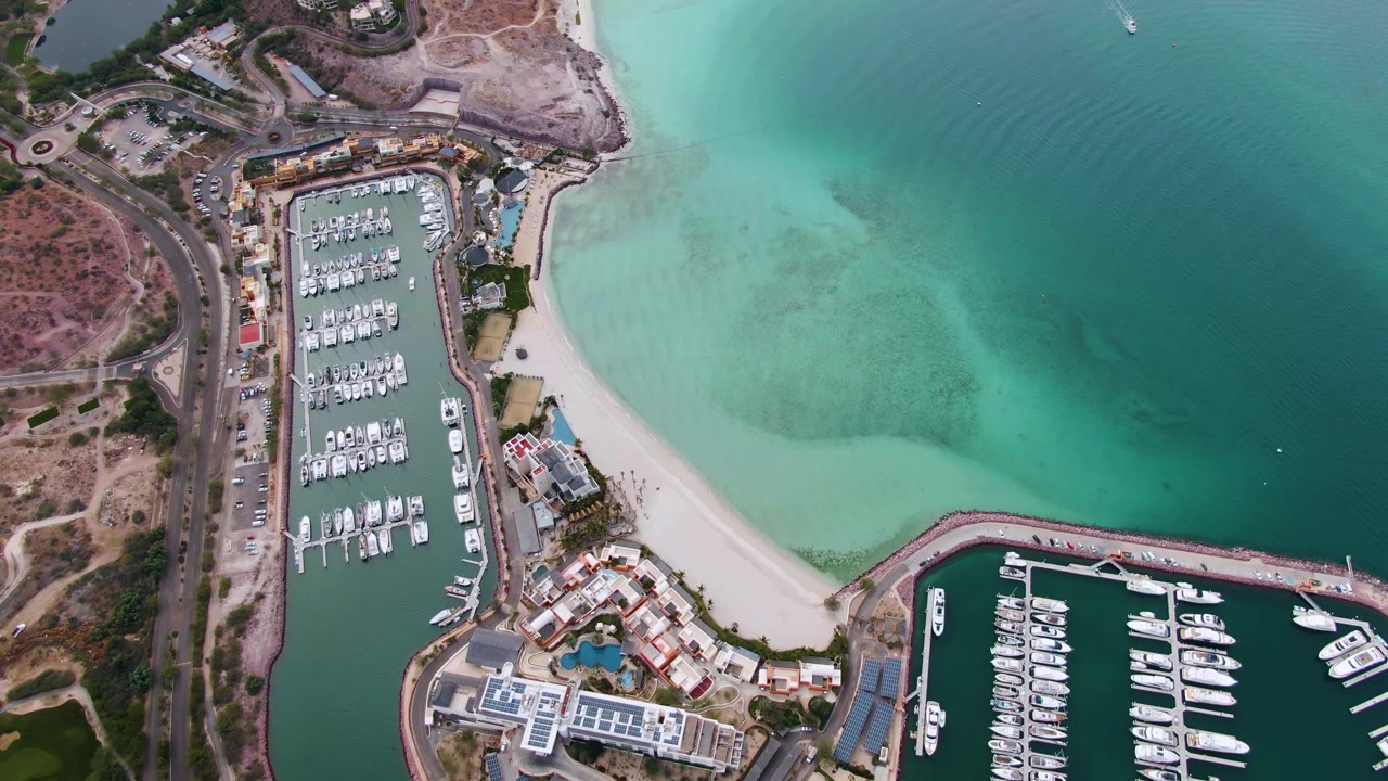 Overhead frame of a La Paz, Baja California Sur marina with rows of boats, a pale curved beach, turquoise coastal water, shoreline roads, and no visible people.