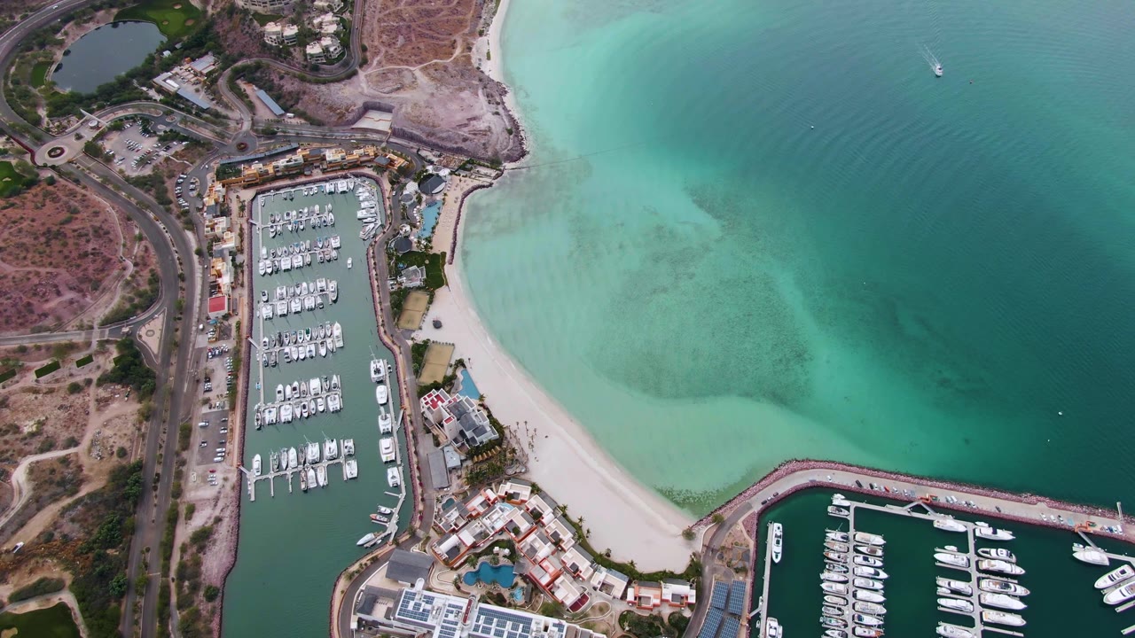 High overhead view of the La Paz marina with boats lined along slips, a curved sandy shoreline, darker open water to the right, and no visible people.