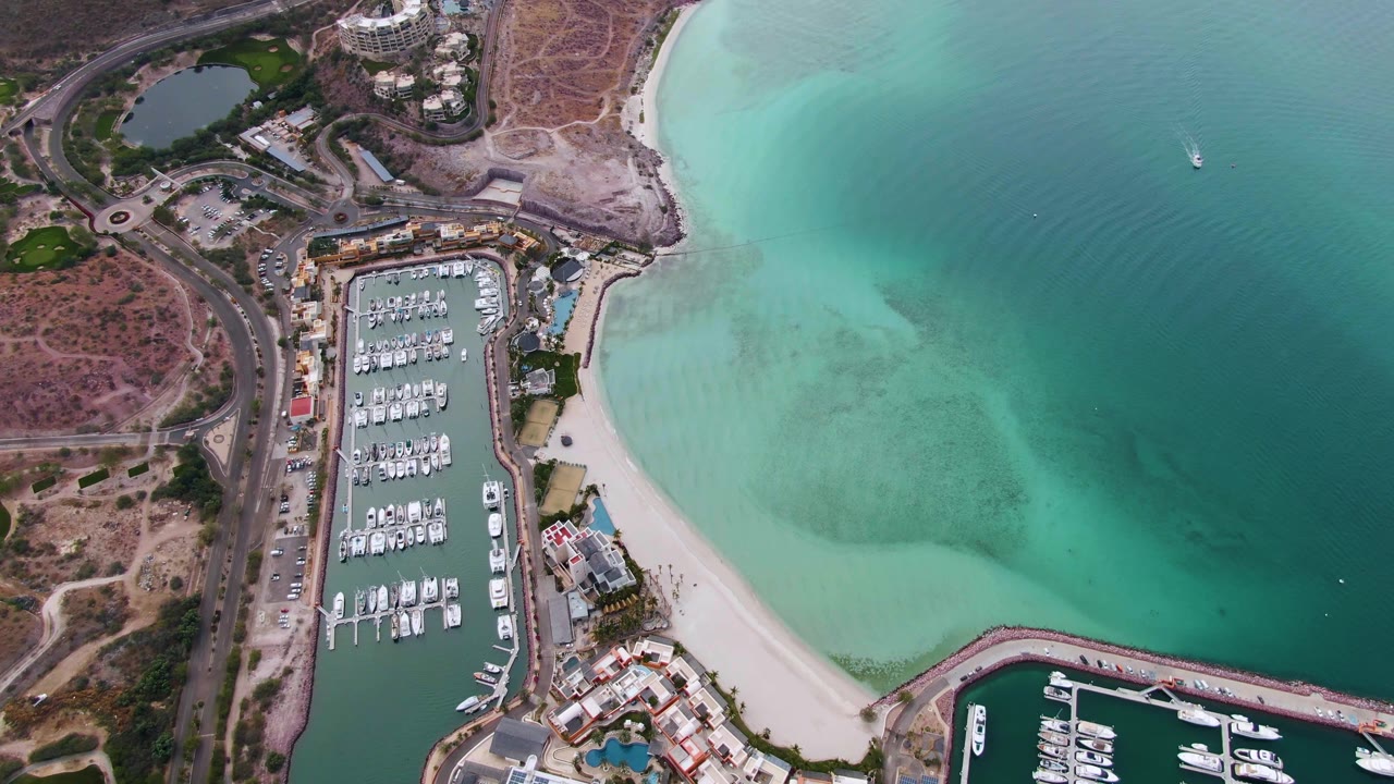 Aerial frame over La Paz, Baja California Sur showing a boat-filled marina beside a pale sandy beach and turquoise water, with dry coastal land and no visible people.