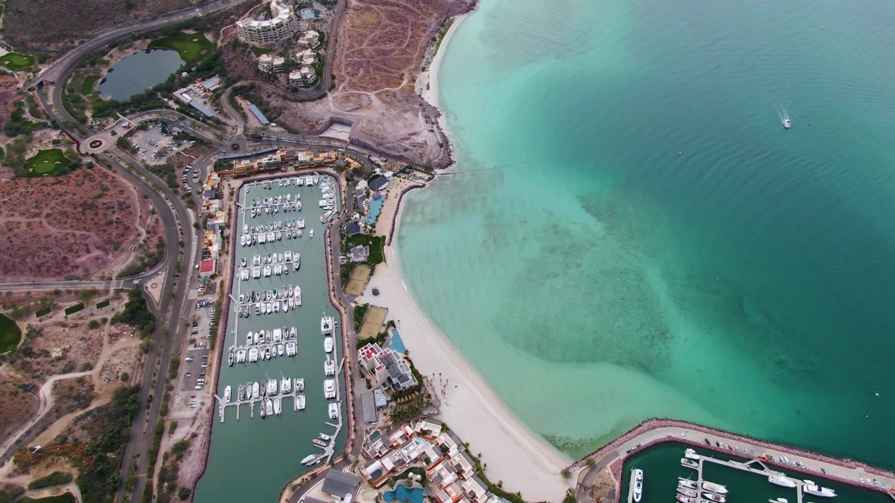 High drone frame over La Paz showing the marina to the left, a coastal road and dry land behind it, a white sand shoreline, clear turquoise water, and no visible people.