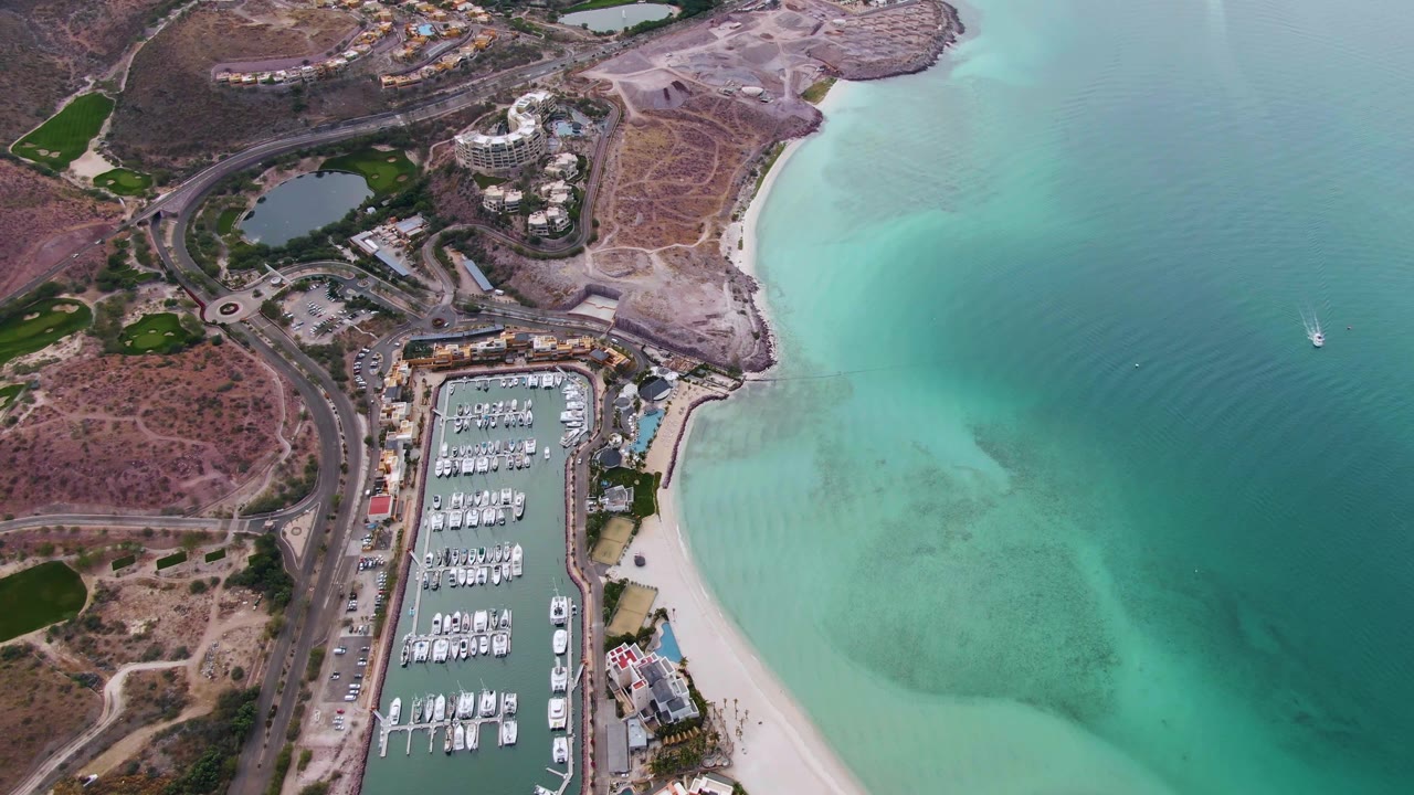 Aerial frame over La Paz with docked boats in the marina, pale sand along the shore, turquoise water filling the right side, a small sailboat offshore, and no visible people.