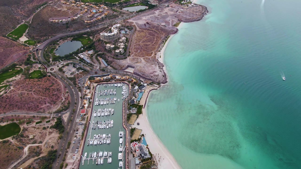 Aerial frame in La Paz, Baja California Sur with the marina centered low in frame, curving roads and dry terrain to the left, turquoise water to the right, and no visible people.